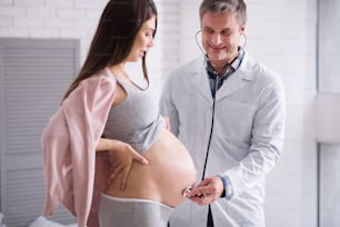 Using instrument. Attractive pregnant woman putting hands on the back and bowing head while looking downwards