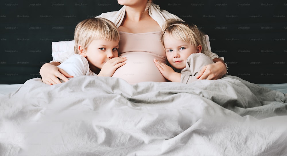 Pregnant woman with her children relaxing in bed. Loving mother and toddlers together at home. Little kids hold hands on pregnant belly of mom. Third pregnancy. Maternity, family, parenting concept.