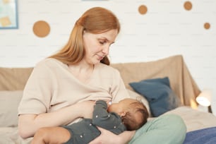 Retrato de tonos cálidos de madre adulta feliz amamantando a un bebé de raza mixta mientras está sentado en la cama en casa, espacio de copia
