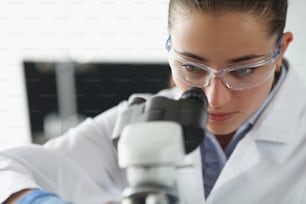 Retrato de una química que investiga la muestra bajo el equipo del microscopio en el laboratorio. Mujer científica explora material, uniforme protector. Concepto de laboratorio