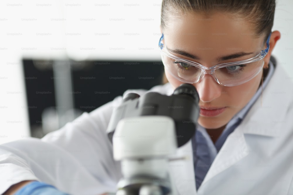 Ritratto di chimico femminile indaga il campione sotto l'attrezzatura del microscopio in laboratorio. La scienziata esplora il materiale, l'uniforme protettiva. Concetto di laboratorio