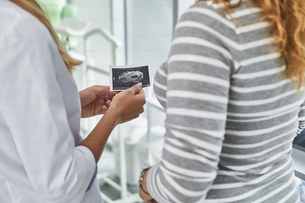 Retrato recortado de la vista trasera del ginecólogo con bata blanca de laboratorio que muestra la ecografía del bebé a la mujer pelirroja