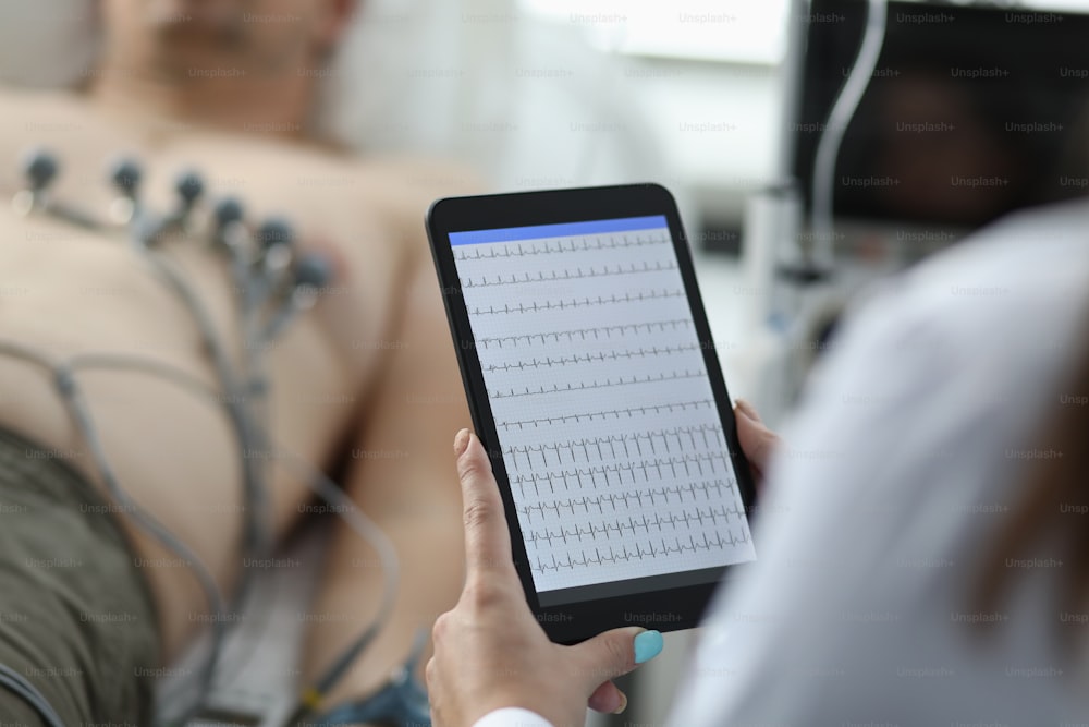 Close-up of female cardiologist holding tablet and examine patients heartbeat. Person laying on hospital bed with special equipment on chest. Medicine and cardiology concept