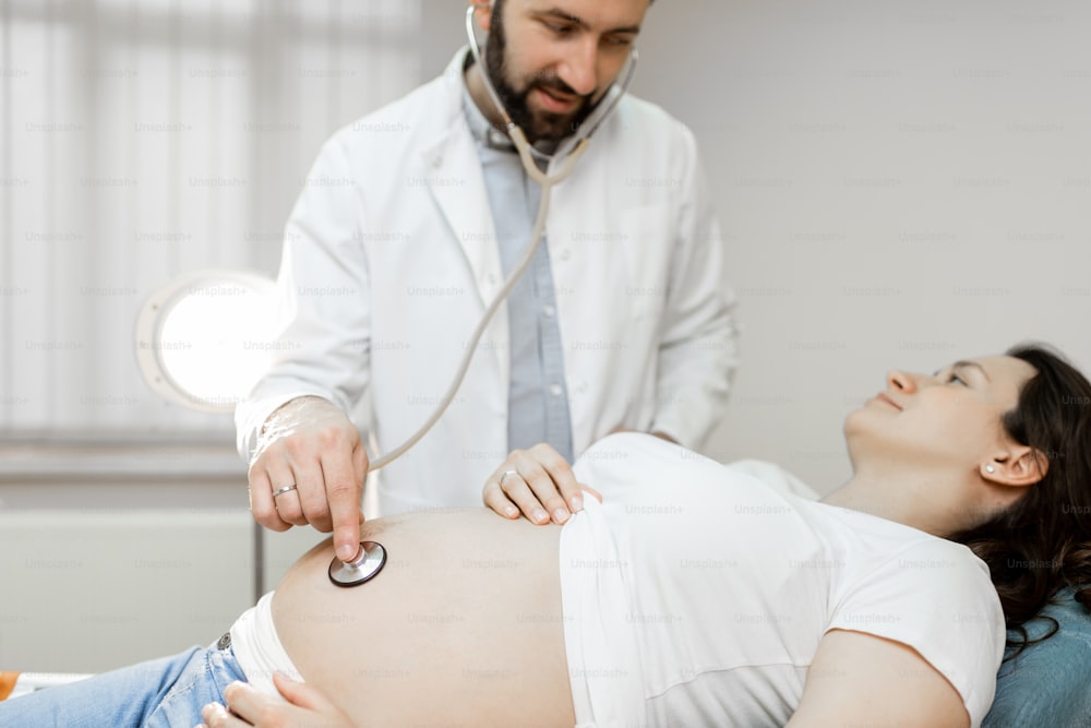 Arzt, der den Bauch einer schwangeren Frau während einer medizinischen Untersuchung mit einem Stethoskop abhört. Konzept der medizinischen Versorgung und Gesundheit während der Schwangerschaft