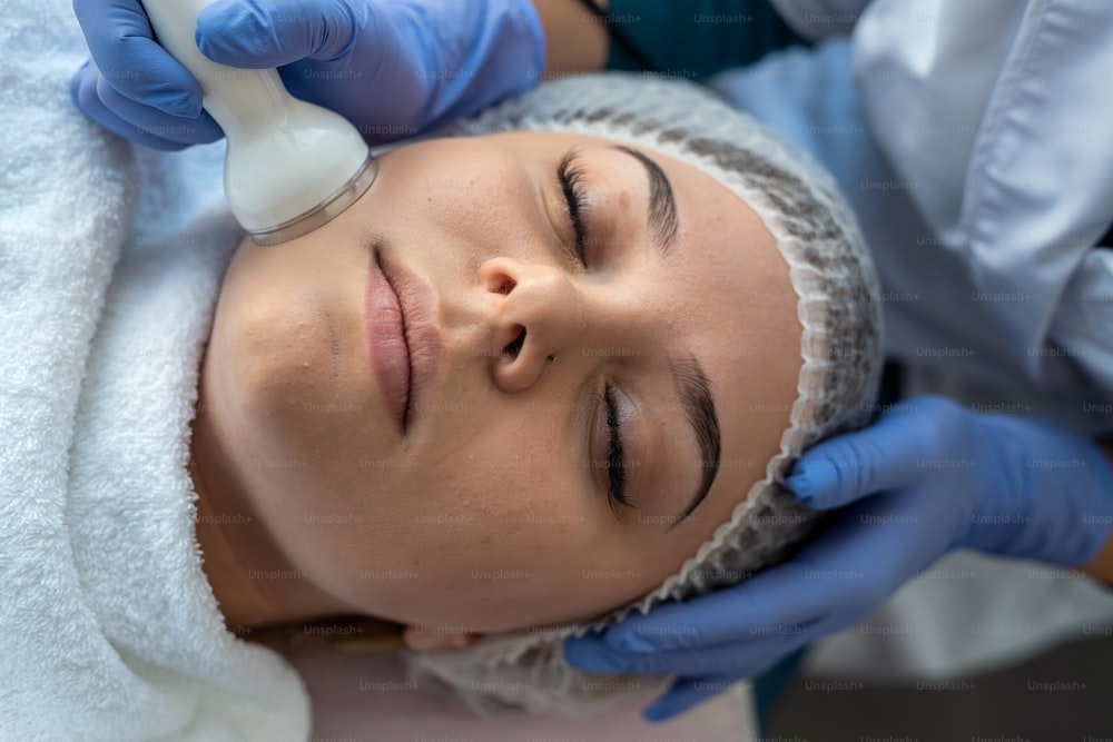 Giovane donna che riceve la procedura professionale cavitazione ad ultrasuoni della pelle facciale come esfoliazione, ringiovanimento e idratazione dal cosmetologo