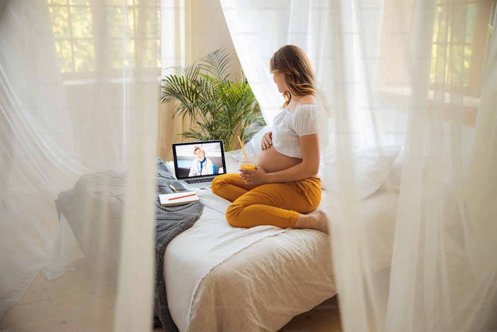 Charming expectant mother sitting on bed and talking with gynecologist through video call stock photo
