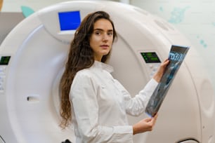 The young women doctor in medical gown looks in camera. Girl with x-ray picture of patient brain in hands. Patient after computer tomography. CT diagnostic in medical clinic