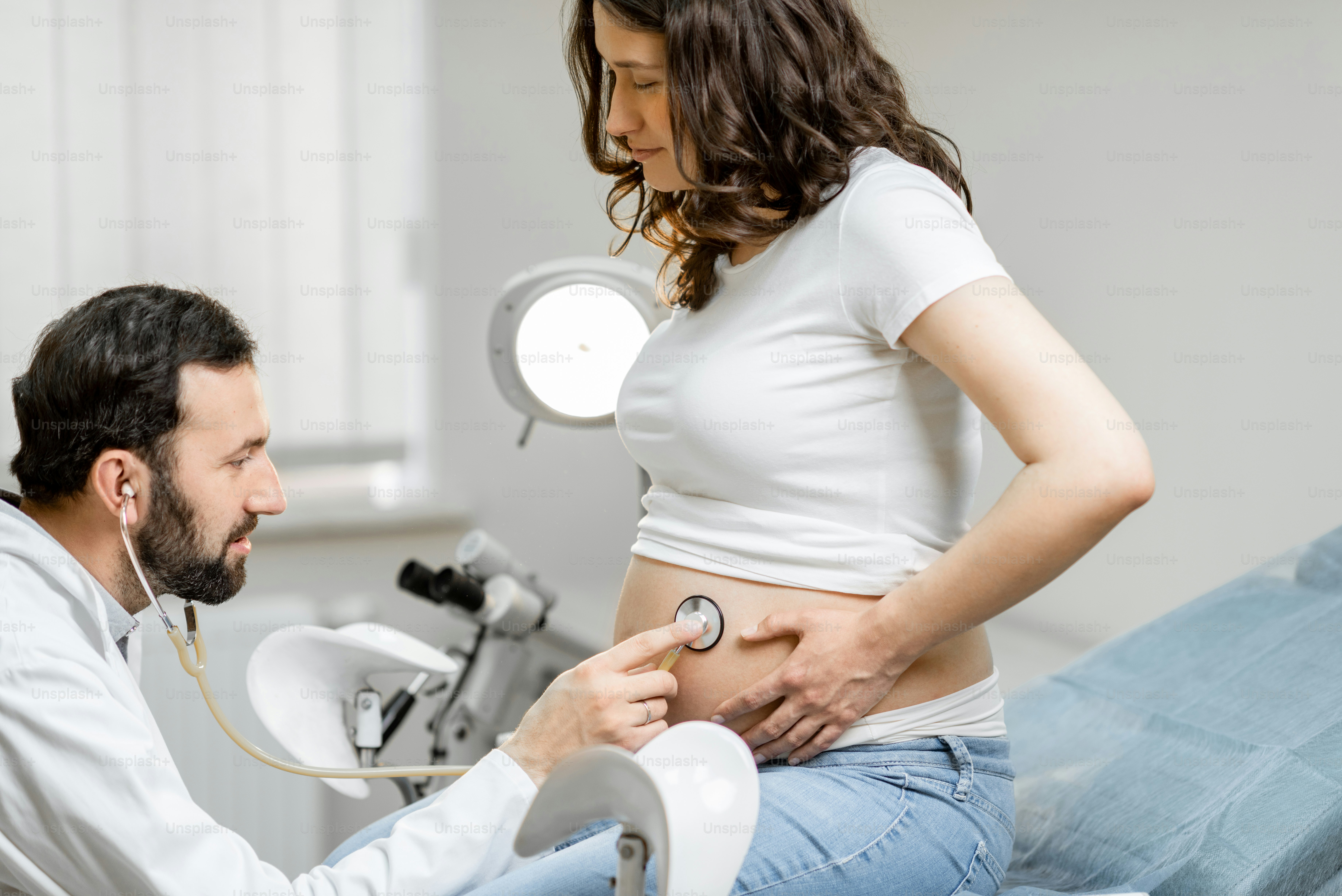 Male doctor listening to a pregnant woman's belly with a stethoscope during a medical examination in the office. Concept of medical care and health during a pregnancy