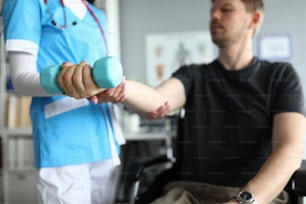 Close-up of professional medical worker helping patient work out arm. Male sitting in wheelchair. Doctor physician in uniform. Modern medicine and disabled people concept