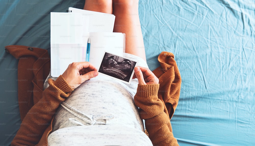 Pregnant woman makes notes, looking at ultrasound image and medical documents. Concept of pregnancy, health care, gynecology, medicine. Mother waiting of baby. Close-up, copy space, indoors.