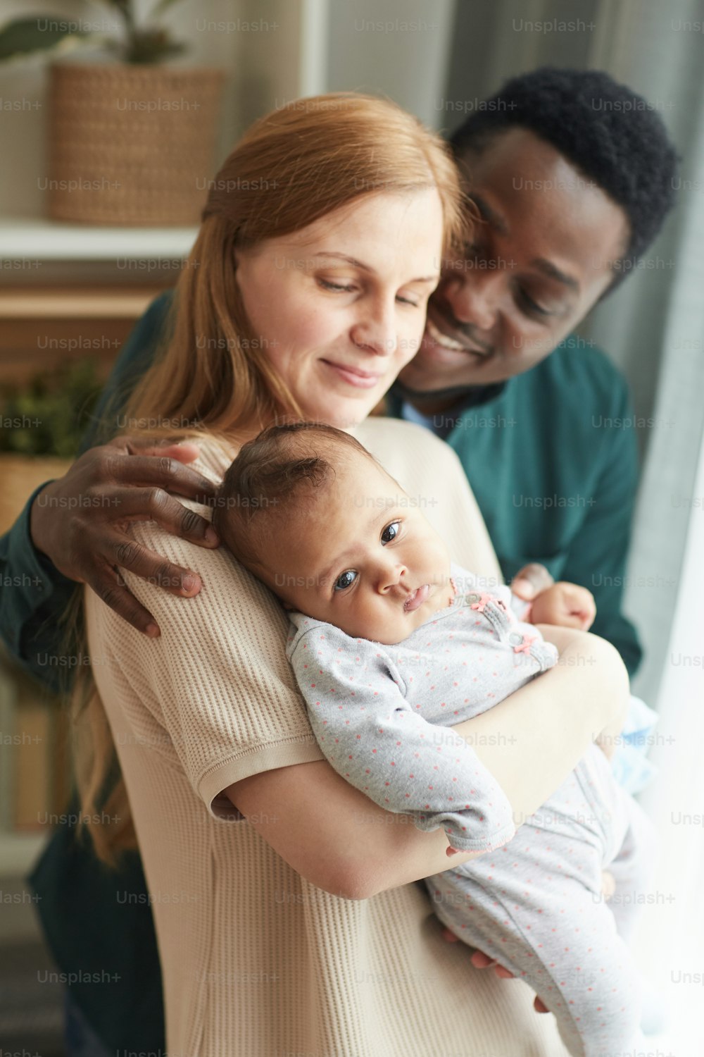 Ritratto verticale di giovane famiglia interrazziale che tiene in mano un bambino carino di razza mista mentre si abbraccia amorevolmente in piedi vicino alla finestra di casa