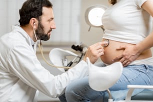 Male doctor listening to a pregnant woman's belly with a stethoscope during a medical examination in the office. Concept of medical care and health during a pregnancy