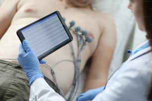 Close-up of female doctor cardiologist check patient results on modern tablet. Protective uniform and gloves. Physician woman diagnostic mans heart. Medicine and ecg concept