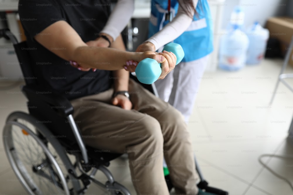 Paralyzed man sit in wheelchair and hold blue dumbbell on his outstretched arm. Femail doctor help patient to knead his hand in clinic.