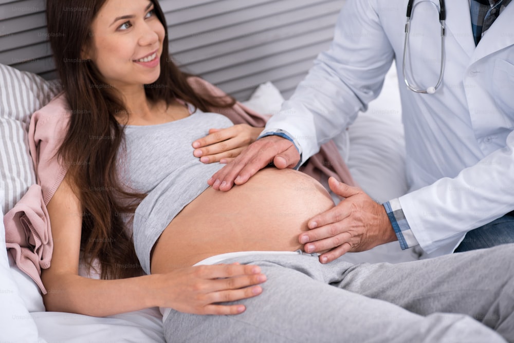 Good form. Delighted brunette keeping smile on her face and looking at doctor while showing her baby bump