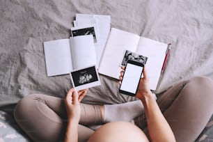 Pregnant woman holding in hands smartphone with empty white display. Close-up pregnant belly of expectant mother with ultrasound photos and medical test reports. Concept of pregnancy, gynecology.