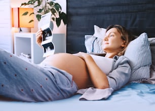 Pregnant woman looking at ultrasound image relaxing in bed at home interior. Expectant mother with pregnant belly waiting and preparing for baby. Concept of pregnancy, gynecologic, healthcare.