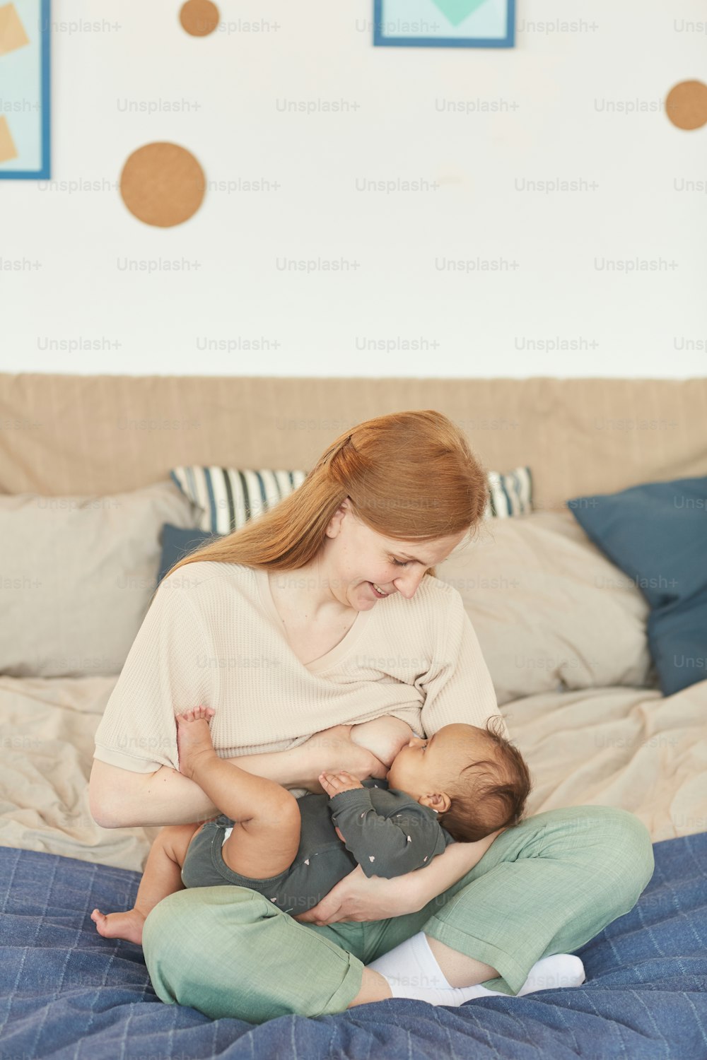 Vertical full length portrait of smiling adult mother breastfeeding mixed-race baby while sitting on bed at home, copy space