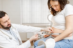 Male doctor measuring pregnant woman's belly with a tape during a medical examination in the office. Concept of medical care and health during a pregnancy