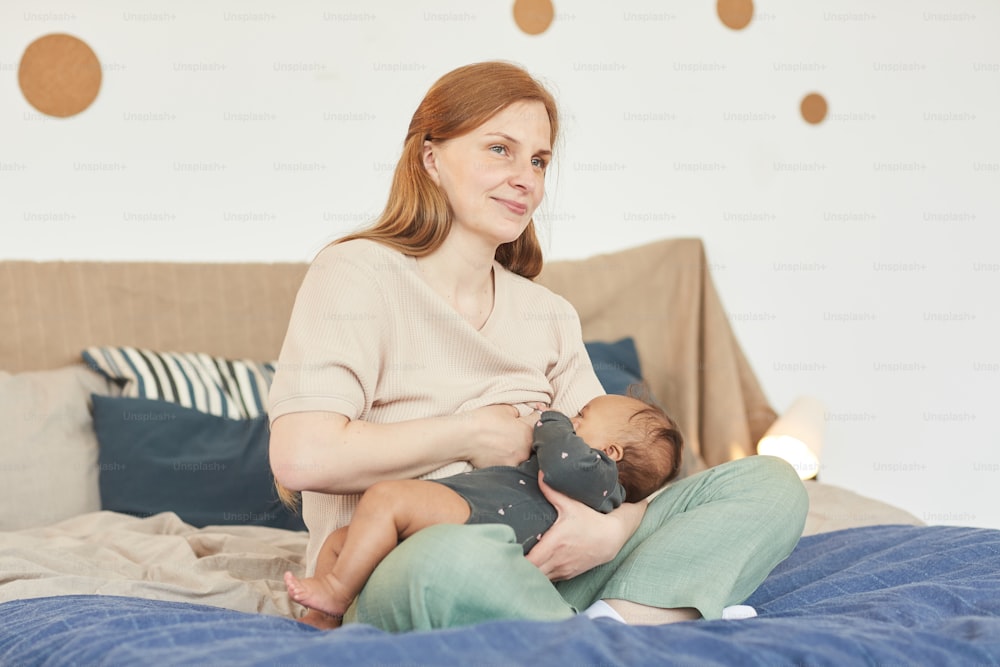 Warm-toned full length portrait of smiling adult mother breastfeeding mixed-race baby while sitting on bed at home, copy space