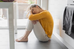Depressed 30 years woman sitting on kitchen floor with hugging knees. Loneliness, sadness. Mental health, drug abuse and social issue concept. Bed news, overworked