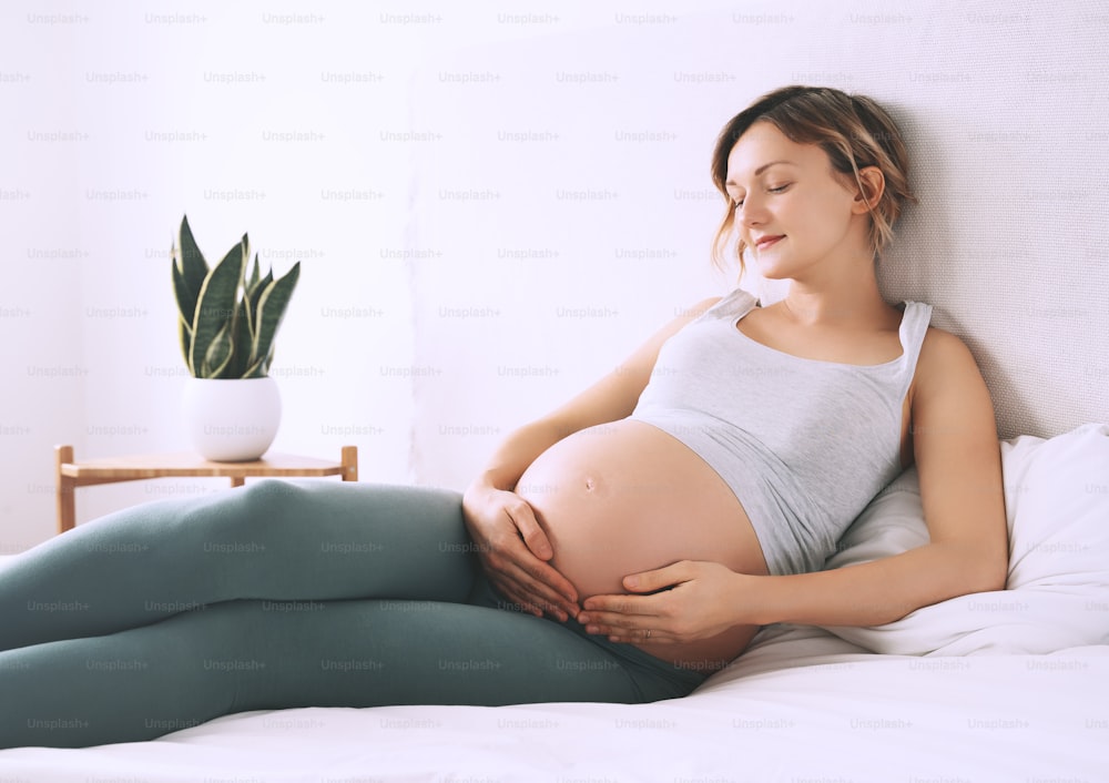 Beautiful relaxed pregnant woman holds hands on her belly in bedroom at home. Young loving mother waiting of a baby. Concept of wellbeing during pregnancy, maternity, healthcare, gynecology.