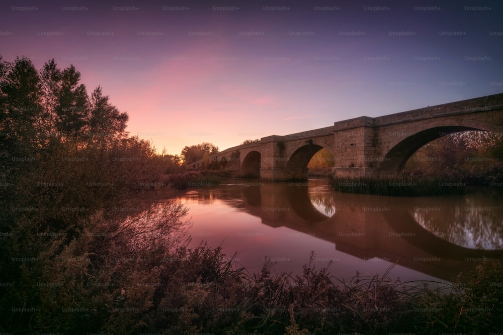 Amazing Landscape. Medieval Bridge over a tranquil river at beautiful colorful sunset.