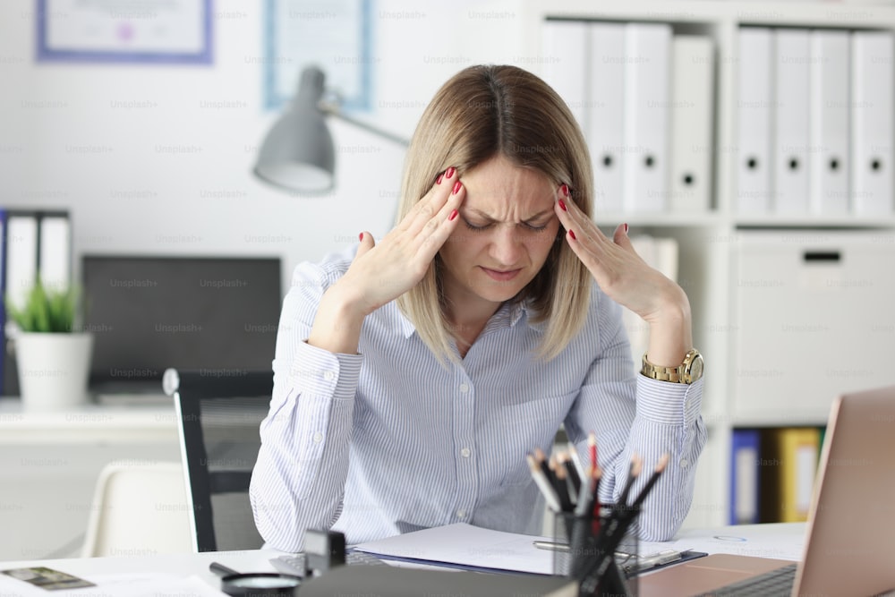 Müde Frau hält sich am Arbeitstisch die Finger an die Schläfen. Konzept für unregelmäßige Arbeitstage