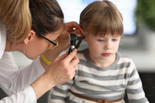 Woman otorhinolaryngologist examines little girl's ear with otoscope in clinic. Diagnostics and treatment of diseases of ear, throat, nose in pediatric practice concept.