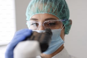 Pesquisador usando máscara de proteção e luvas olha através do microscópio. Pesquisa e desenvolvimento de novos conceitos de vacinas