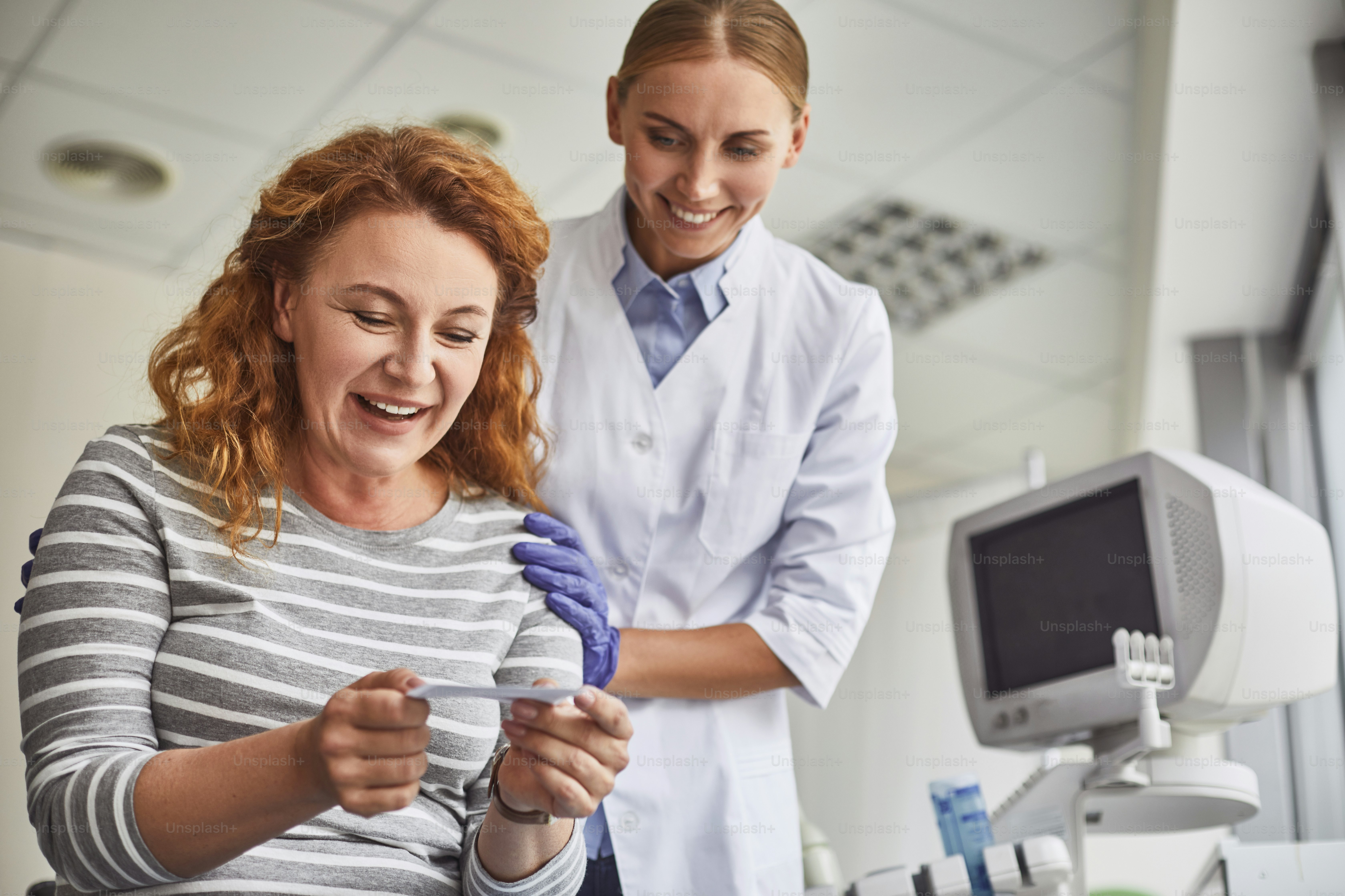 I am pregnant. Waist up portrait of joyful mature lady looking at baby sonogram while smiling doctor touching her shoulder