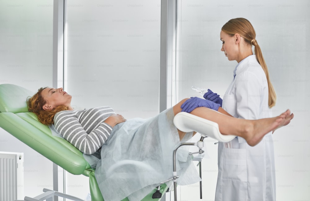 Side view portrait of young gynecologist in white lab coat and sterile gloves holding medical vaginal speculum. Red-haired woman with closed eyes lying in gynecological chair