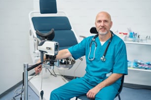 Serious gynecologist is sitting in a medical cabinet while his hand rested on a detail of a chair for gynecological manipulations