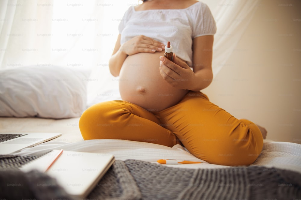 Expectant mother sitting on bed with laptop and holding bottle of serum stock photo