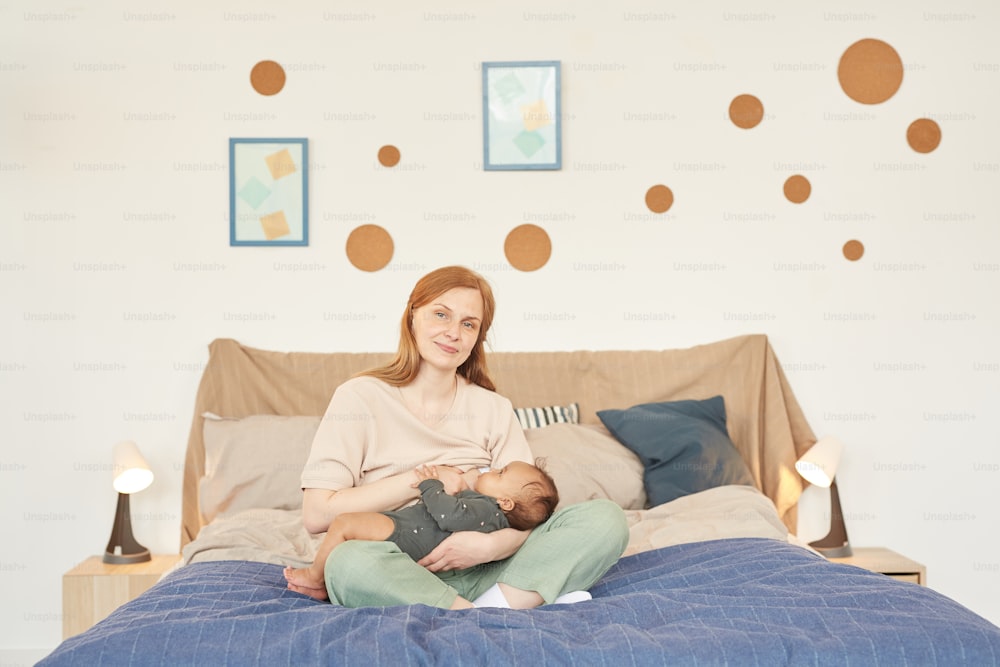 Warm-toned full length portrait of happy adult mother breastfeeding mixed-race baby and looking at camera while sitting on bed at home, copy space