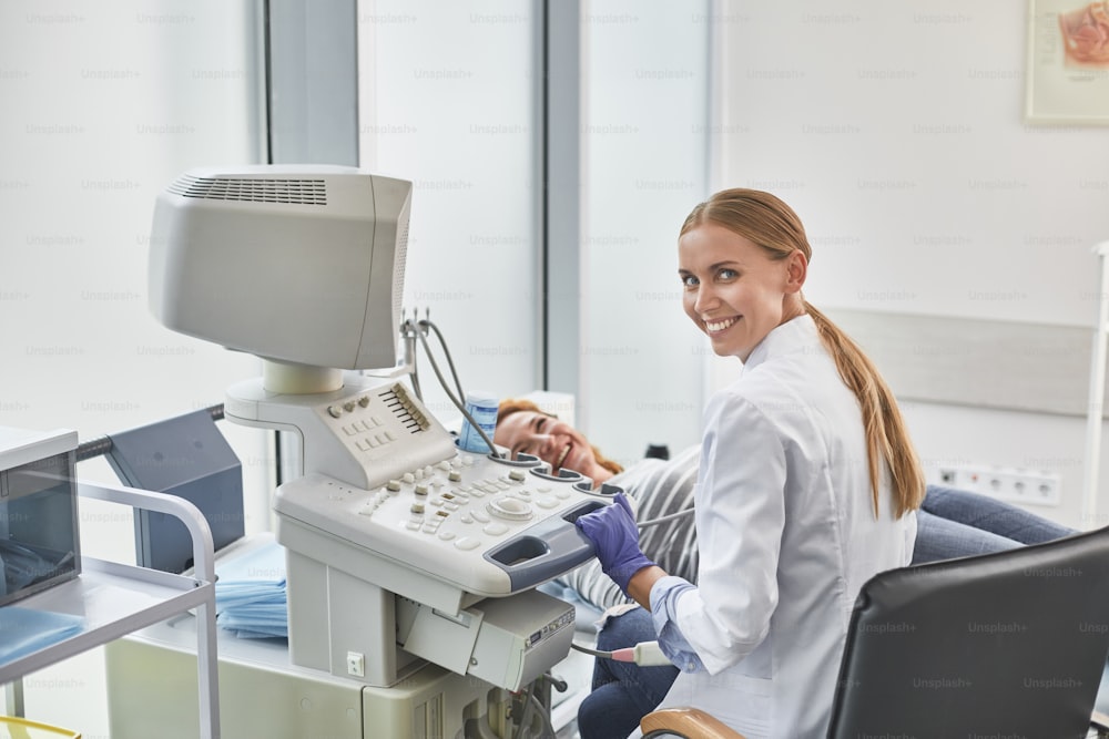 Back view portrait of gynecologist in white lab coat and sterile gloves using ultrasound scanner during medical examination of her patient. Smiling red-haired woman lying on daybed
