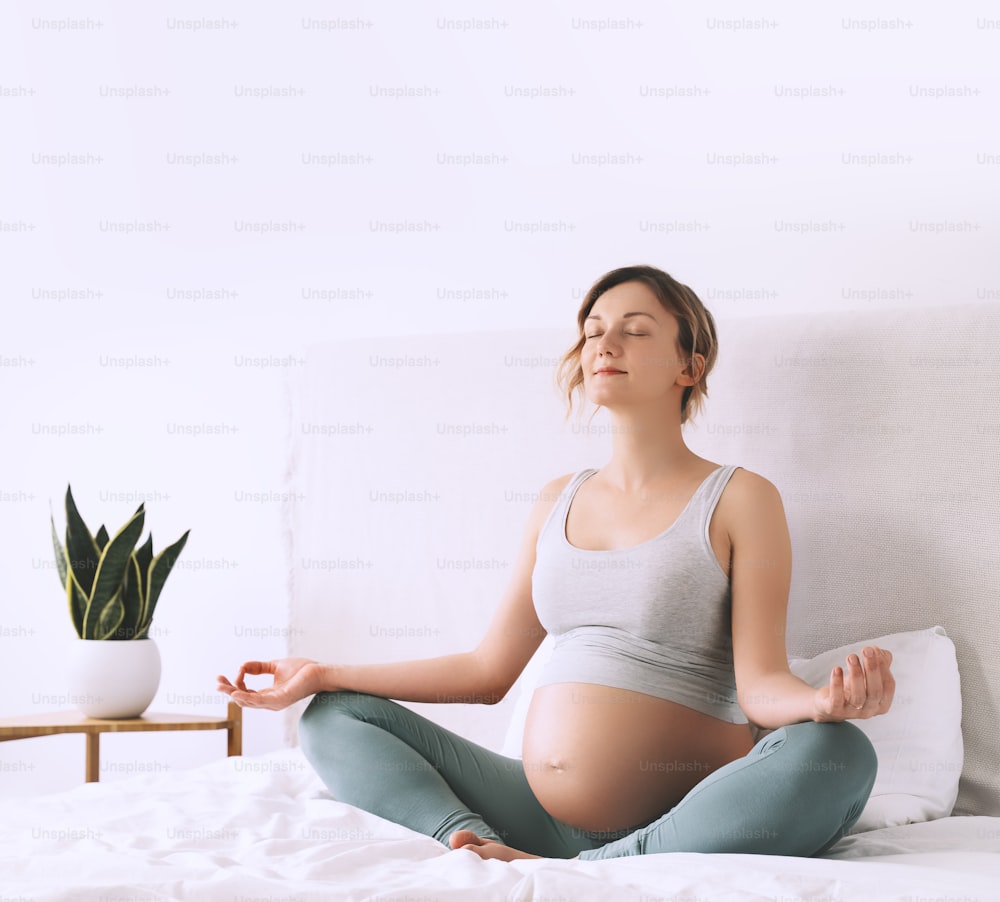 Pregnant woman in lotus pose doing meditation or breathing exercises for healthy pregnancy and preparing body for childbirth. Young expectant mother practicing yoga at home.