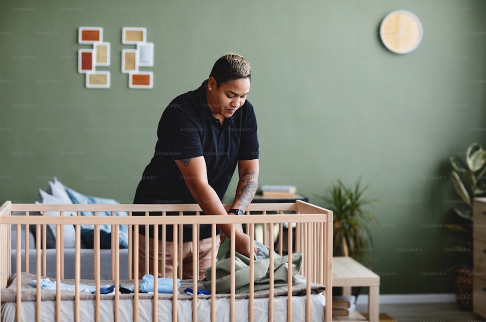 Portrait of tattooed gay woman preparing baby crib in minimal home interior, copy space