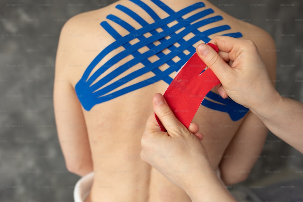 Closeup cropped doctor, physiotherapist hands taping blue and red stripes on back muscles for Kinesio, kinesiology therapy treatment. Female patient injured, backache, pain suffer after training