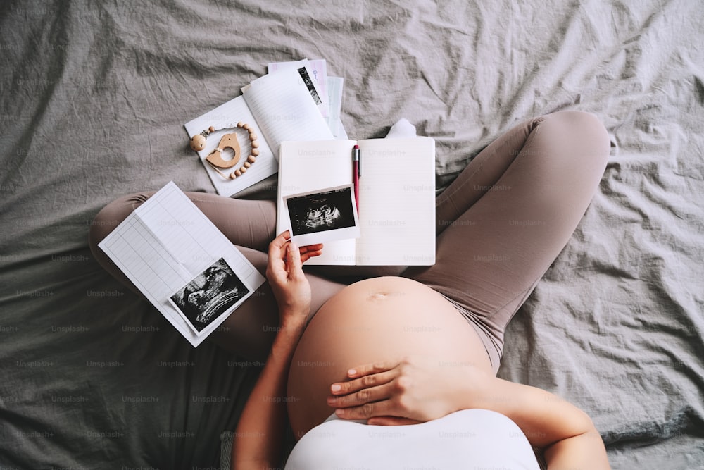 Pregnant woman holding ultrasound image looking at notebook and medical test reports. Expectant mother with pregnant belly waiting for baby. Concept of pregnancy, gynecologic, healthcare.