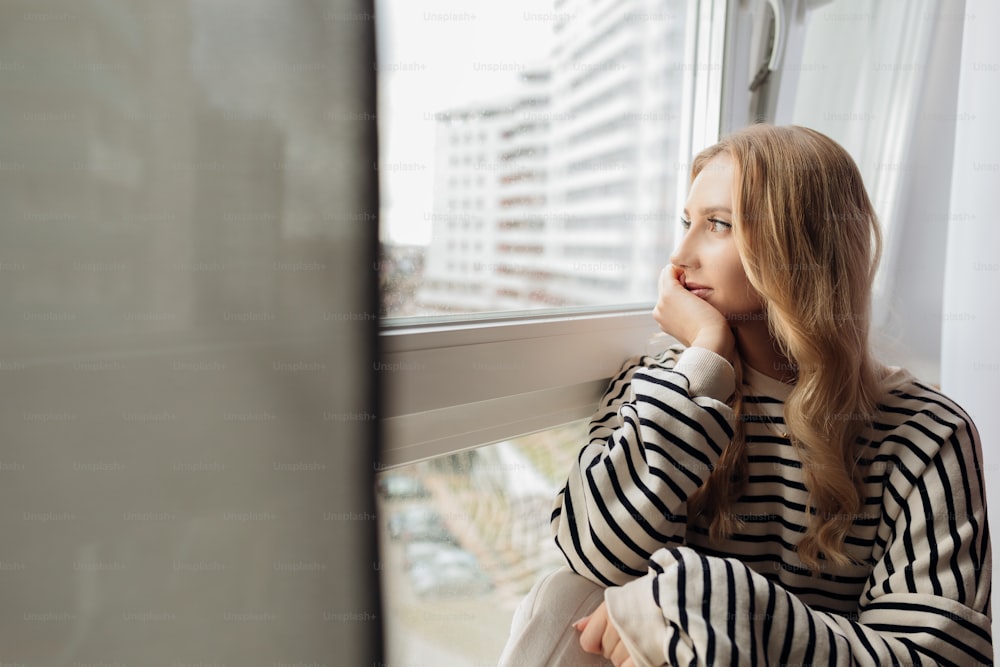 Portrait of pensive thoughtful blond pretty dreaming depressed woman sitting on window sill looking distance street. Lonely and tired mood, isolation. Light day. Broken up boyfriend, end relationship