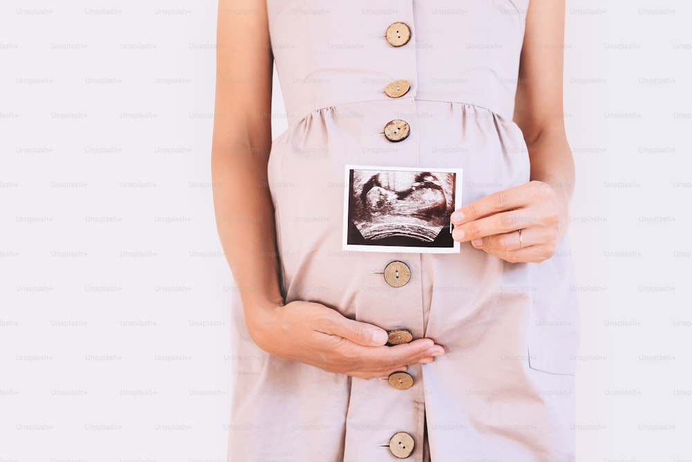 Pregnant woman holding ultrasound baby image. Close-up of pregnant belly and sonogram photo in hands of mother. Concept of pregnancy, gynecology, medical test, maternal health.