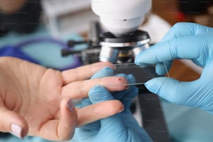 Close-up of laboratory worker taking blood from female finger for investigation. Modern microscope on table in lab. Medicine, chemistry, science concept