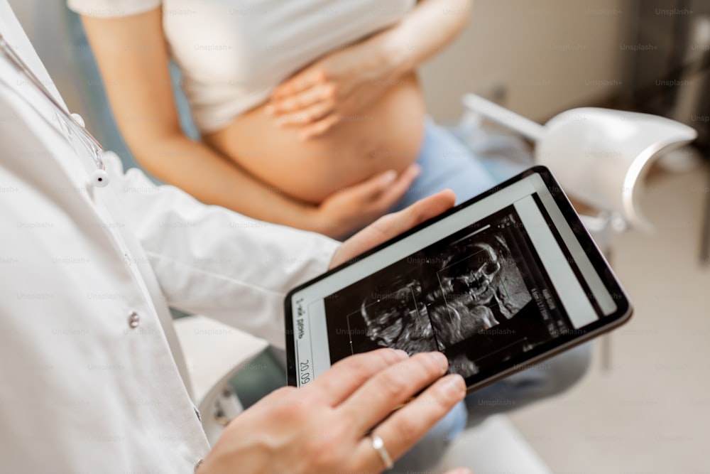 doctor with an ultrasound scan of unborn child on a digital tablet during an examination with a pregnant woman in the office, cropped view without faces