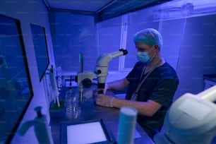 Man in sterile uniform, protective face mask, gloves and hat conducting researches under microscope in laboratory with ultra-violet light