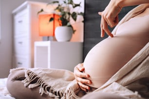 Beautiful pregnant woman holds hands on her belly in bedroom at home. Young loving mother waiting of a baby. Concept of pregnancy, maternity, healthcare, gynecology. Close-up, indoors.