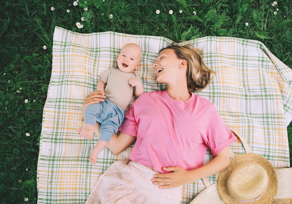 Smiling mom and baby lying on blanket on green grass at summer. Family relaxing and having picnic outdoors. Beautiful mother with her baby on nature. Concept of motherhood, human happiness, eco life.