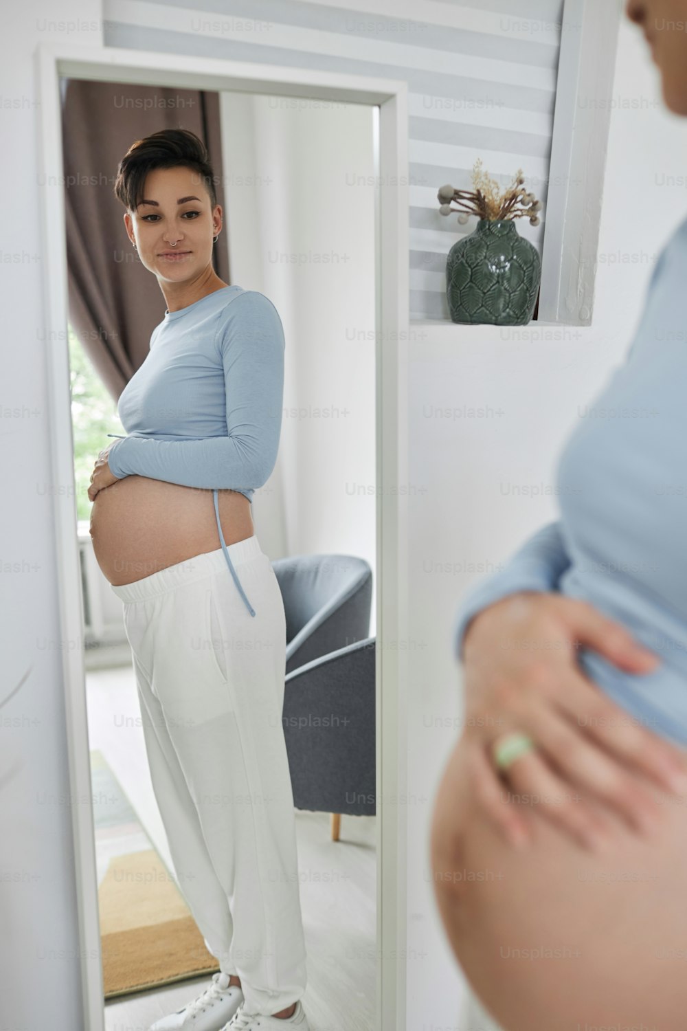 Side view portrait of young pregnant woman looking in mirror and measuring baby bump with tape