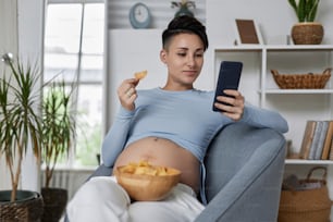 Porträt einer modernen schwangeren Frau, die ein Smartphone benutzt und Chips isst, während sie sich zu Hause entspannt