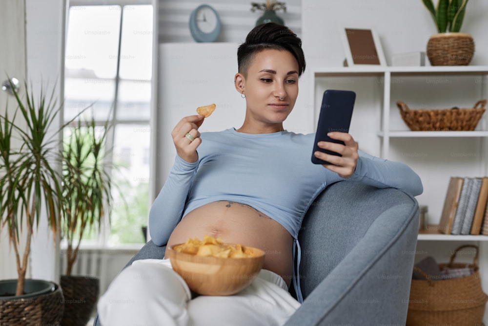 Portrait of modern pregnant woman using smartphone and eating chips while relaxing at home